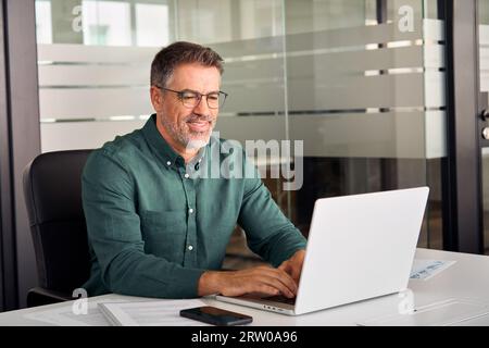 Un uomo d'affari anziano e sorridente che lavora con un computer portatile seduto alla scrivania. Foto Stock