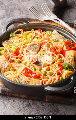 Spaghetti con pesce bianco, peperone, piselli verdi, aglio e pomodori da vicino in una padella sul tavolo. Verticale Foto Stock