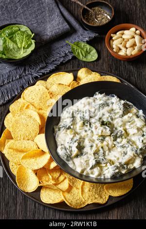 salsa cremosa di carciofi spinaci e mandorle con patatine fritte in recipiente su piatto nero su tavola di legno scuro con ingredienti, vista verticale dall'alto Foto Stock