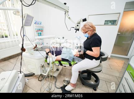 Vista laterale delle dentiste sedute sulla sedia, dentatrici che fissano i denti. Ispezione medica, esame del problema, tenuta di attrezzature, indossare uniformi, guanti e maschera. Concetto di odontoiatria. Foto Stock
