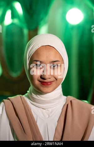 Una donna musulmana con un velo bianco e vestiti bianchi sorridente molto carina senza trucco sul viso in una stanza verde di notte Foto Stock