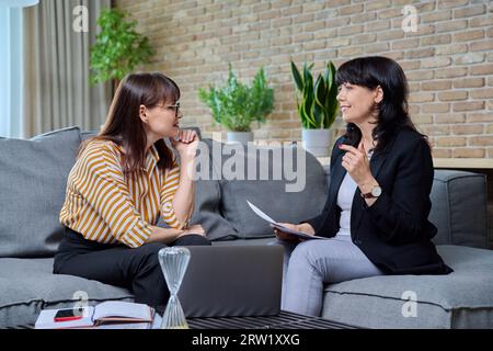 Due donne mature d'affari sedute sul divano in ufficio, a parlare, a parlare di lavoro Foto Stock