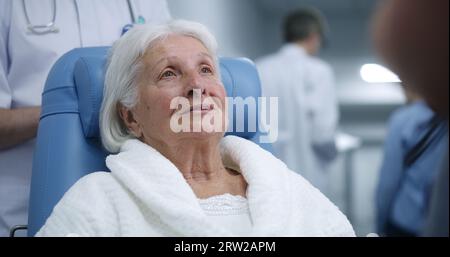 Donna anziana in sedia a rotelle di trasferimento comunica con il medico nel corridoio della clinica. La paziente femminile parla con il medico prima della terapia. Personale medico e pazienti che camminano in background nel corridoio dell'ospedale. Foto Stock