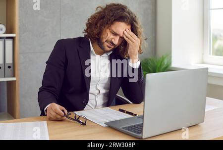 Uomo stanco che soffre di mal di testa che lavora sul suo posto di lavoro, massaggiando la fronte. Foto Stock