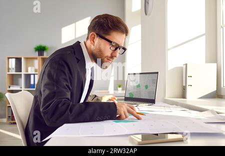 Topografo catastale seduto alla scrivania dell'ufficio e che lavora con i piani della città e le mappe catastali Foto Stock