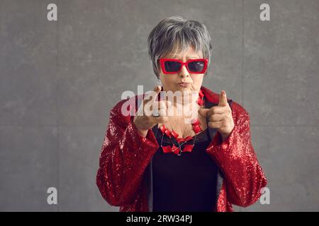 Donna anziana con giacca da discoteca fa un volto divertente e punta il dito indice verso la fotocamera Foto Stock