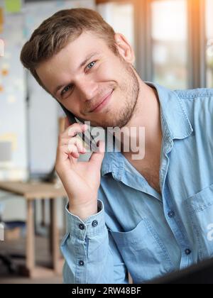 Bell'uomo parla al telefono e sorride. L'operatore risponde alla chiamata telefonica. Il ragazzo caucasico in camicia di denim è seduto in ufficio e parla al telefono cellulare. Emozioni positive. Servizio di supporto cordiale. Foto Stock