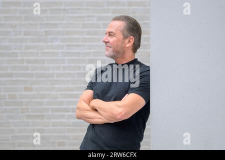 L'attraente uomo dai capelli grigi si appoggia contro un muro con le braccia incrociate e guarda in lontananza davanti a un muro di mattoni bianchi Foto Stock