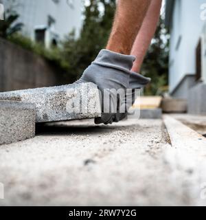 Vista ravvicinata ad angolo basso delle mani maschili in guanti da lavoro protettivi posa di piastrelle di cemento per pavimentare una superficie intorno a una casa. Foto Stock