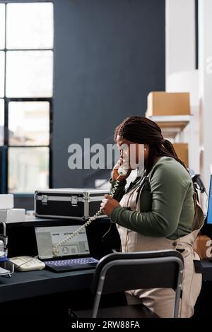 Lavoratore che prepara l'ordine discutendo della detalis di consegna con il cliente remoto al telefono fisso in magazzino. Dipendente afroamericano che analizza la lista di controllo delle merci su un computer portatile nel magazzino Foto Stock