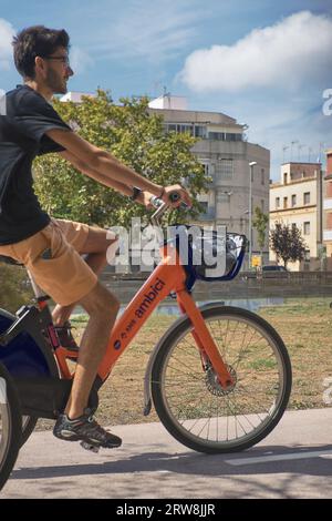 Viladecans, Spagna - 18 settembre 2023: Giovane uomo caucasico in pantaloncini corti e t-shirt sorridente con gli occhiali che cavalcano lungo la pista ciclabile di un parco e di un elec Foto Stock