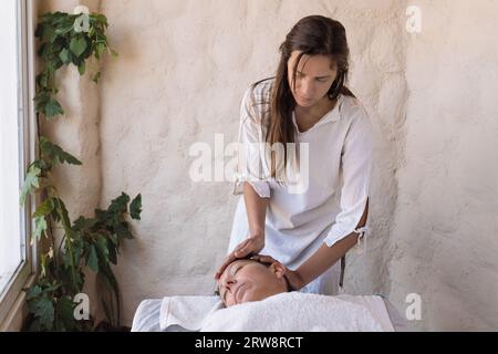 Bellissimo giovane terapeuta che esegue massaggi nel salone della spa. Ritratto frontale dell'estetista che fa un massaggio facciale a una rilassata cliente femminile. bea facciale Foto Stock