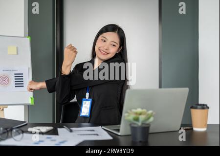 Una felice e bella donna d'affari asiatica si è allungata le braccia dopo aver finito il lavoro su un laptop alla sua scrivania, sentendosi spensierata e a suo agio Foto Stock