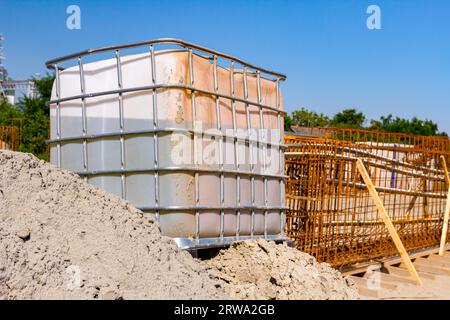 Serbatoio dell'acqua quadrato bianco, cubico, con griglia metallica, enorme serbatoio per liquido su pallet di legno in cantiere, sullo sfondo è rinforzato scheletro del ponte Foto Stock