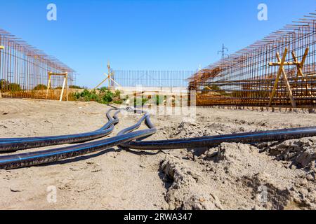 I lunghi tubi dell'acqua in plastica nera con striscia blu sono collocati a terra in cantiere sullo sfondo si trova lo scheletro del ponte in costruzione. Foto Stock