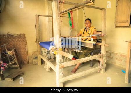 Srimongal, Bangladesh, circa luglio 2012: Donna a piedi nudi dai capelli neri con sciarpa gialla siede su una sedia di legno e si intreccia con un panno blu a Srimongal Foto Stock