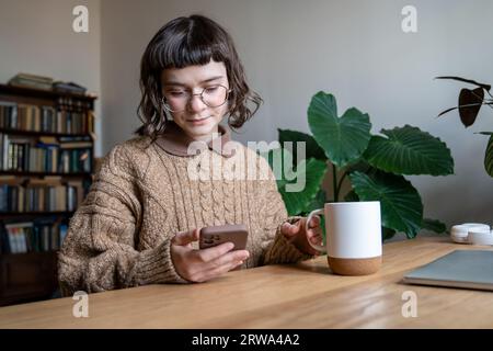 Una studentessa beve tè, usa il telefono. Una donna nella biblioteca del college con il telefono si prende una pausa dalle lezioni Foto Stock