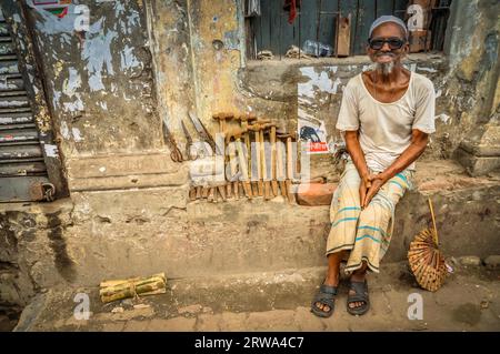 Dhaka, Bangladesh, circa luglio 2012: Un vecchio sorridente con la barba bianca siede su una panchina di pietra e vende martelli e forbici per strada a Dacca Foto Stock
