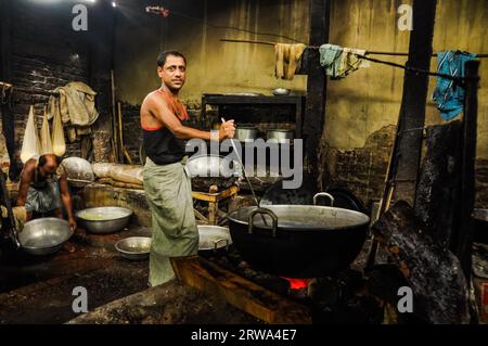 Chittagong, Bangladesh, circa luglio 2012: L'uomo nativo dai capelli neri tiene un grande mestolo e mescola il cibo in pentola sopra il fuoco in cucina nel villaggio rurale vicino Foto Stock