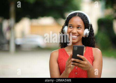 Ritratto frontale di una donna di colore felice che indossa cuffie e tiene il telefono che guarda lateralmente ascoltando musica Foto Stock