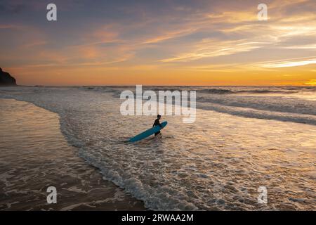 Fai un giro in barca sull'oceano per fare un giro al tramonto. Il surfista cammina sulla spiaggia al tramonto, pronto per una sessione serale. Surfboarder che cammina tra le onde dell'oceano Foto Stock