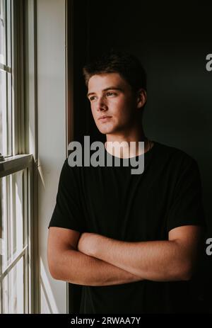 Ritratto di un bel ragazzo adolescente che guarda fuori dalla finestra della stanza buia. Foto Stock