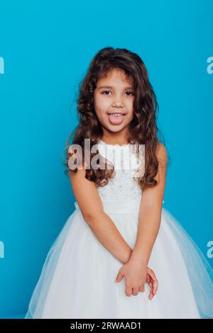 una bambina con un vestito bianco mostra la sua lingua su uno sfondo blu Foto Stock