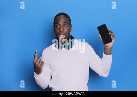 Ritratto di un giovane ragazzo afro-americano che indossa un maglione bianco casual in posa isolato su sfondo blu. Giovane uomo millenario spensierato che tiene Foto Stock