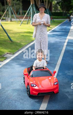 Un simpatico bambino multirazziale cavalca un'auto rossa con telecomando mentre sua madre cammina dietro di lui guidando un veicolo giocattolo Foto Stock