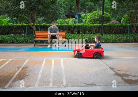Un simpatico bambino multirazziale cavalca un'auto rossa con telecomando mentre suo padre è seduto sulla panchina e guida un veicolo giocattolo Foto Stock
