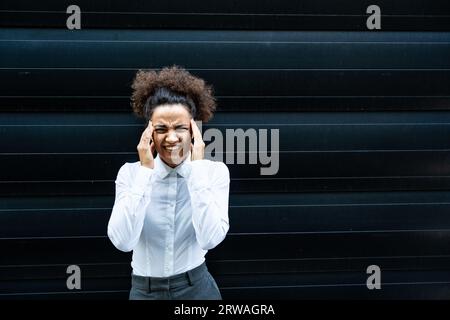 Donna stressata sta impazzendo tirandosi i capelli in frustrazione da vicino di una giovane donna d'affari. Lavoratrice d'ufficio in piedi contro la parete nera Foto Stock