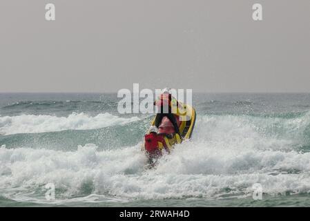 Membro del bagnino RNLI sulla moto d'acqua con un altro su una slitta di salvataggio che si dirige verso il mare spietato al largo di Perranporth Beach, Cornovaglia, Regno Unito. Foto Stock