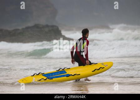 Membro del bagnino RNLI con bordo di salvataggio da surf in direzione di mari irregolari al largo di Perranporth Beach, Cornovaglia, Regno Unito. Foto Stock