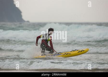 Membro del bagnino RNLI con bordo di salvataggio da surf in direzione di mari irregolari al largo di Perranporth Beach, Cornovaglia, Regno Unito. Foto Stock