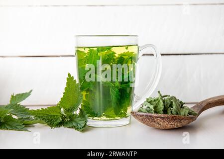 Tè alle erbe a base di Urtica dioica secca, nota come ortica comune, bruciare l'ortica o pungere foglie di ortica in una tazza di vetro trasparente. Sfondo in legno bianco. Foto Stock