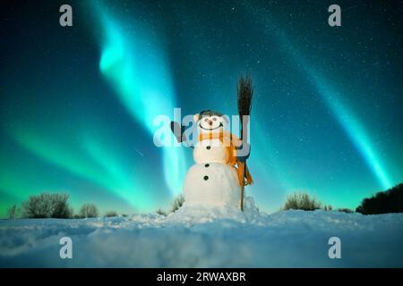 Divertente pupazzo di neve in elegante cappello rosso e cuoio capelluto rosso sul campo nevoso sullo sfondo di un incredibile cielo stellato con Aurora borealis. Panorama notturno incredibile. Aurora boreale in campo invernale Foto Stock