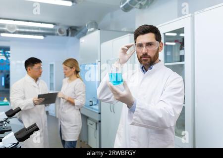 Ritratto di un serio chimico maschio, scienziato in un laboratorio e con in mano un matraccio con una sostanza blu per la ricerca. I suoi colleghi parlano dietro di lui. Foto Stock