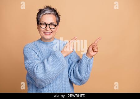 Ritratto fotografico di una graziosa nonna Fingers punta spazio vuoto indossa indumenti in maglia blu alla moda isolati su sfondo di colore beige Foto Stock