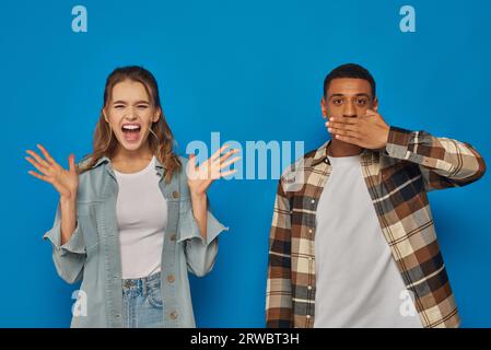 reazione di coppia interrazziale su sfondo blu, donna emotiva che urla vicino a un uomo afro-americano Foto Stock