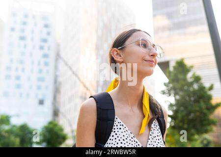 Dal basso di una giovane donna felice con occhiali da vista con zaino vicino a edifici alti e moderni che guardano lontano Foto Stock