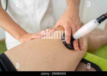 Medico professionale ritagliato non riconoscibile in uniforme utilizzando lo strumento di terapia a onde d'urto sul ginocchio del cliente durante l'appuntamento in clinica moderna Foto Stock