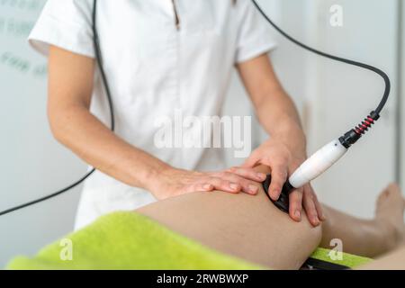 Medico professionista non riconoscibile in uniforme che utilizza lo strumento di terapia a onde d'urto sul ginocchio del cliente durante l'appuntamento in clinica moderna Foto Stock