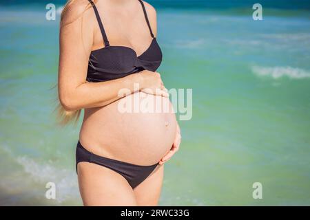Radiosa donna incinta in costume da bagno, sullo sfondo mozzafiato di un mare turchese. Bellezza serena della maternità vicino alla riva Foto Stock