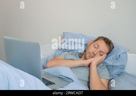 Un giovane uomo caucasico si addormenta sul letto davanti al laptop. Un freelance maschio che dorme dietro un computer portatile a letto la mattina. Uomo designer che lavora in remoto Foto Stock