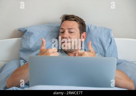 Bell'uomo caucasico sorridente e che mostra il gesto del pollice in alto durante la video chat sul laptop. Un uomo che si trova a letto e che ha una conversazione online la mattina Foto Stock