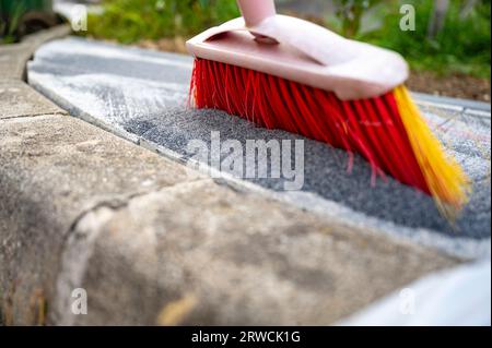 Vista ravvicinata ad angolo basso della scopa rossa brillante che spinge un'apertura piastrellata di sabbia negli spazi di un marciapiede appena pavimentato. Foto Stock