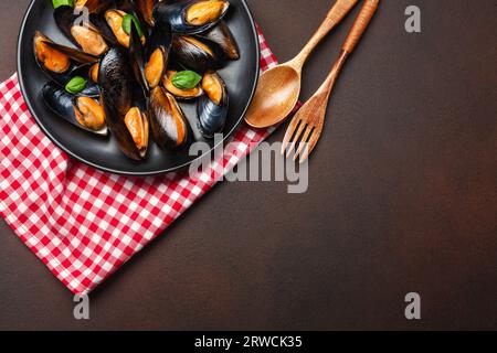 Cozze di mare e foglie di basilico in un piatto nero con asciugamano, cucchiaio di legno e forchetta su fondo arrugginito. Vista dall'alto. Foto Stock