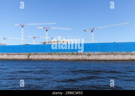 Gru a torre bianche sotto il cielo azzurro, il cantiere si trova dietro la recinzione di metallo blu sulla costa del fiume Neva, San Pietroburgo, Russia Foto Stock