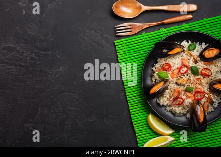 Riso fritto con cozze di pesce, gamberi, basilico in un piatto nero con cucchiaio di legno e forchetta su tappetino di bambù verde e tavola in pietra. Vista dall'alto. Foto Stock