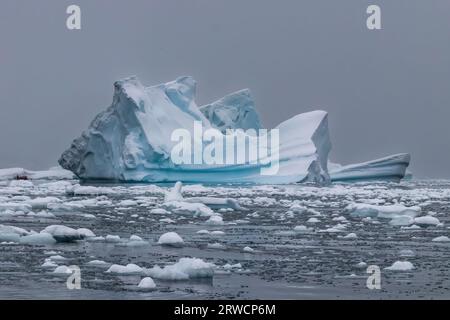 Iceberg con ghiaccio blu e bianco vicino alla penisola antartica. Cielo grigio innevato sullo sfondo; acqua calma in primo piano coperta di ghiaccio. Foto Stock